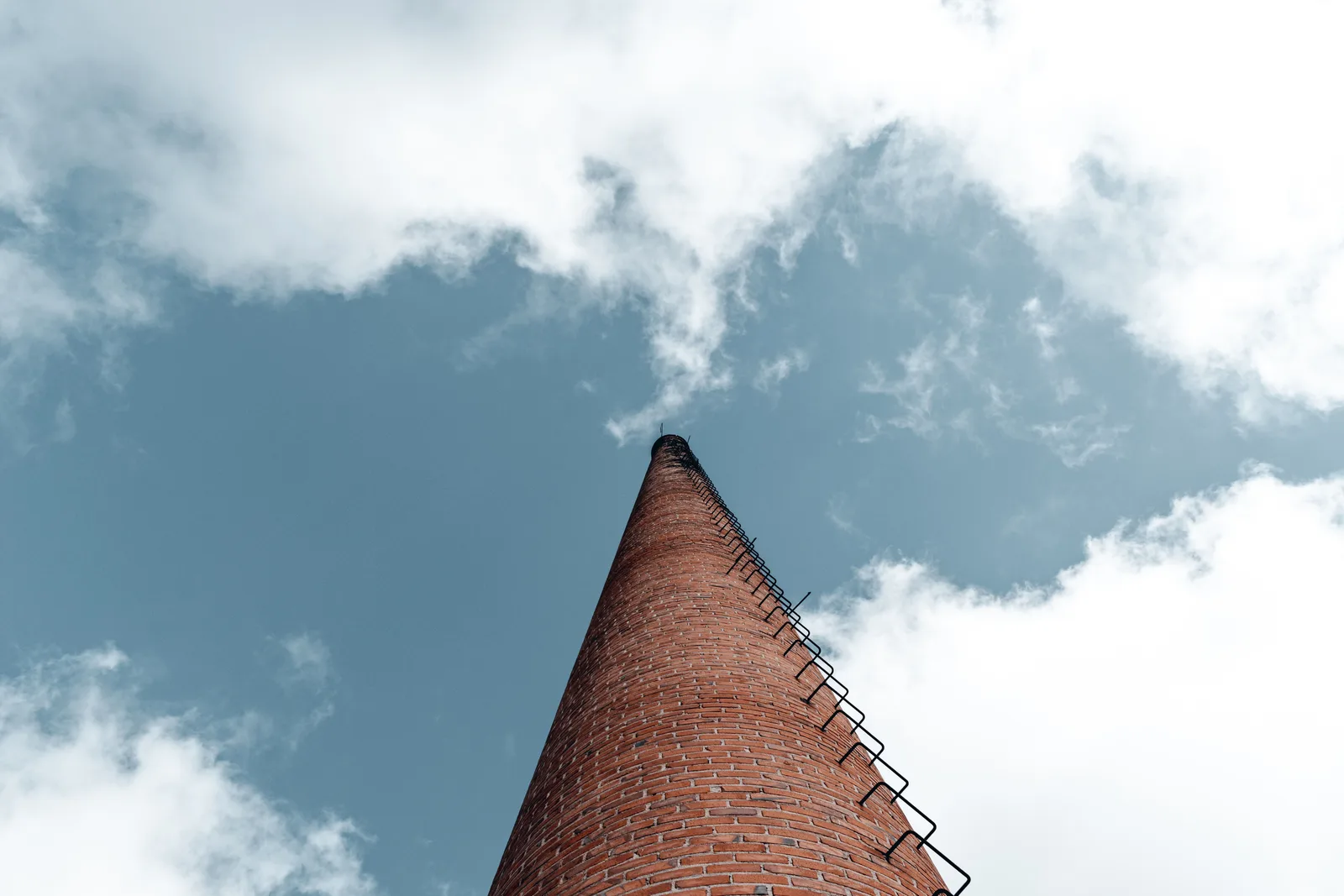 A towering brick chimney with a ladder reaching into a blue sky scattered with white clouds, captured from a low angle. What chimney sweep insurance costs, and what it covers