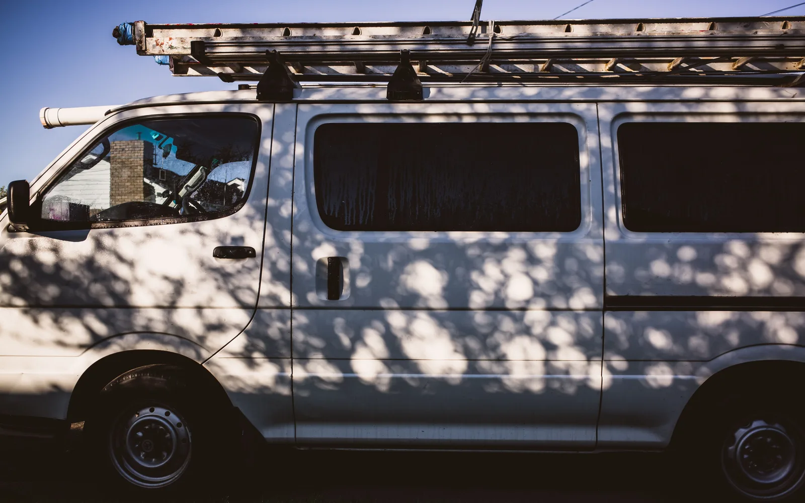 A white van with a ladder on top casting intricate shadows from nearby trees outdoors. Chimney Sweep Van Setup: Compact Van vs Full-Size